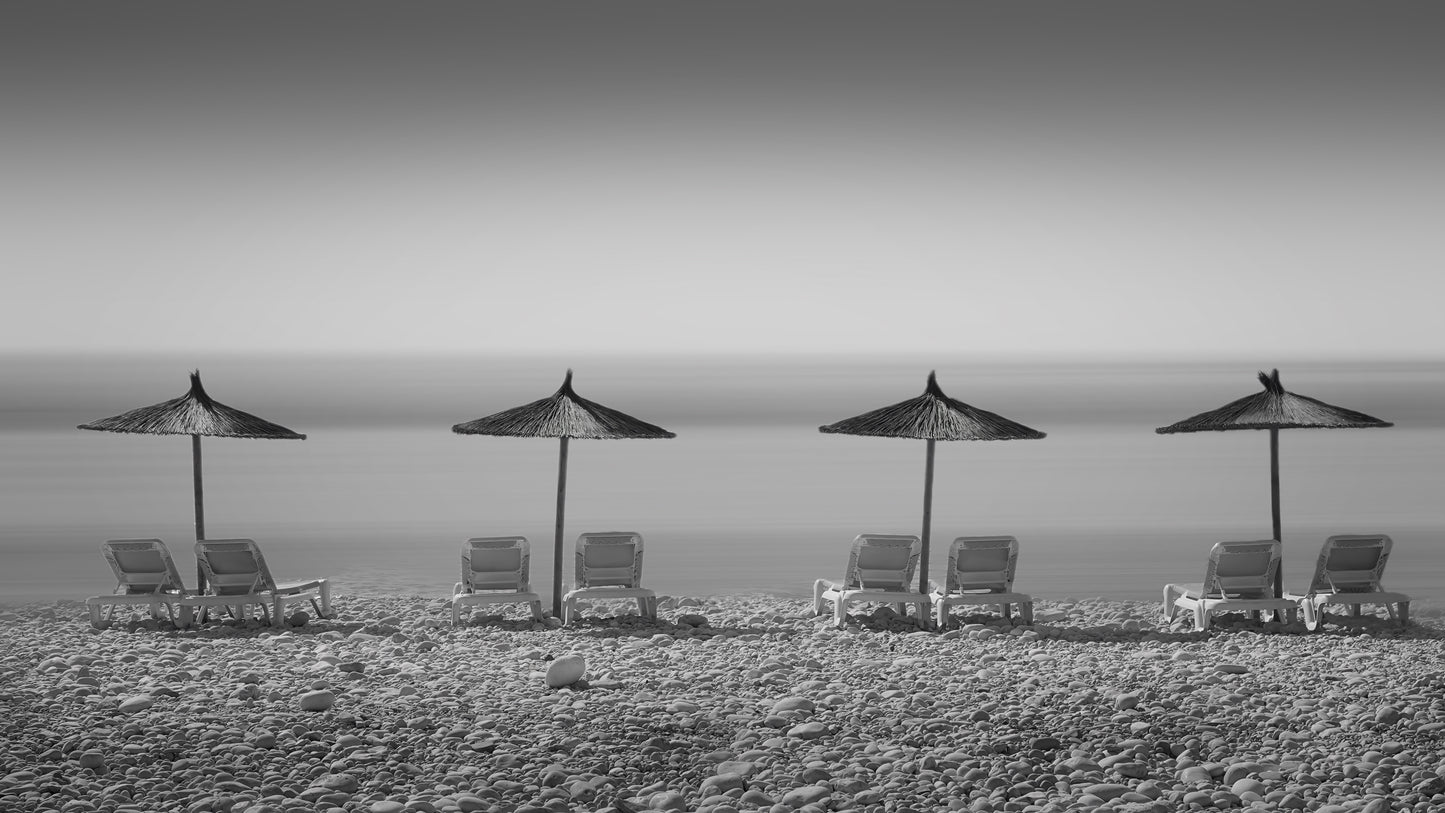 Black and white scene of beach chairs and umbrellas on a sandy beach with ocean in the background.